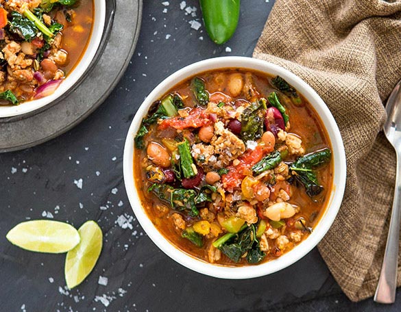 Sausage, bean and vegetable chili in bowls with lime and a burlap napkin on a dark surface.
