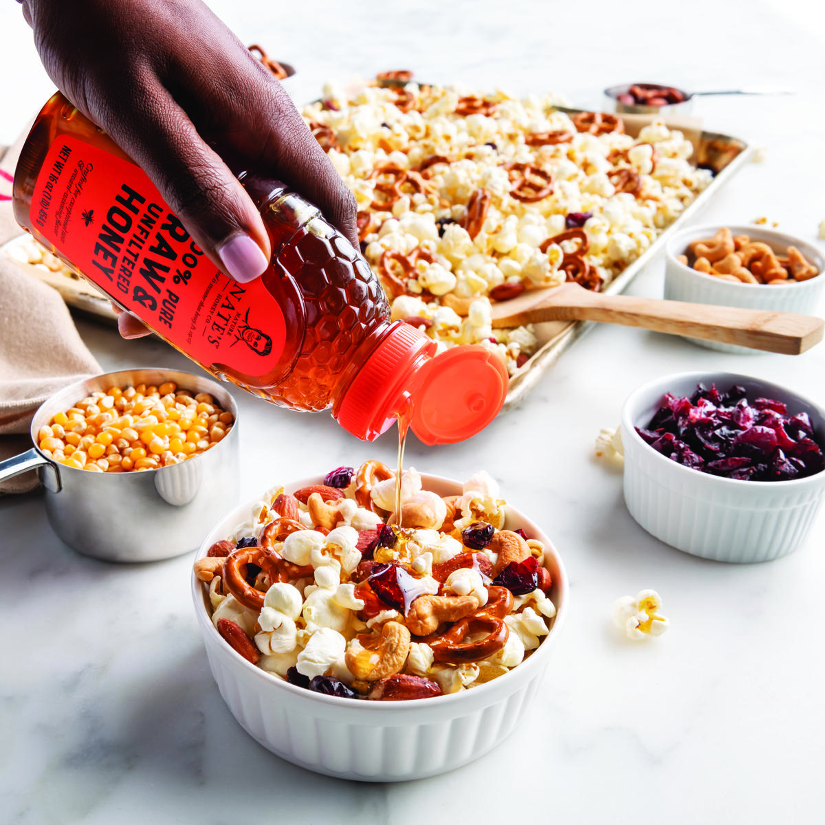 A hand pouring Nate’s honey onto a bowl of nuts, dried fruit and popcorn, with ingredients all around on marble.