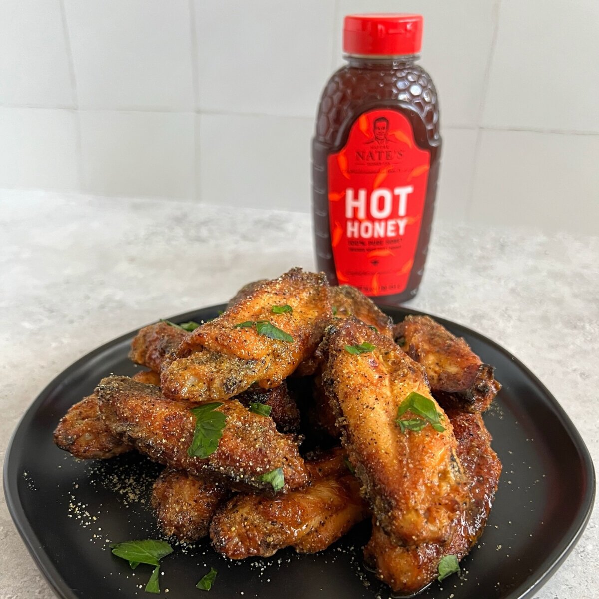 Crispy chicken wings garnished with green herbs and served on a black plate, alongside a bottle of Nate’s hot honey.