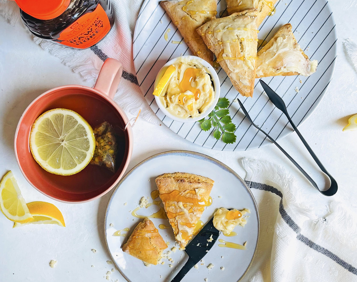 Lemon poppyseed scones drizzled with honey on a plate, a cup of tea, and a bottle of Nate’s honey on a white surface.