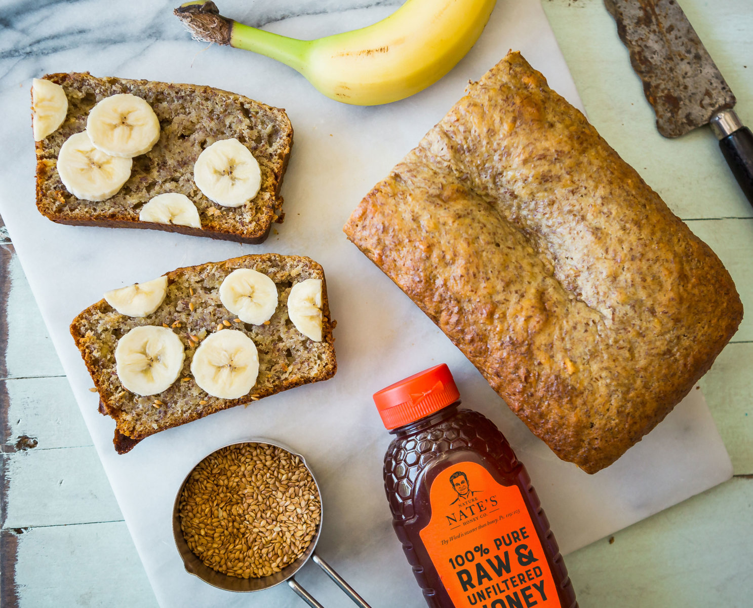 Banana bread topped with slices of banana, a banana, Nate’s honey bottle, flaxseeds and a knife on paper.