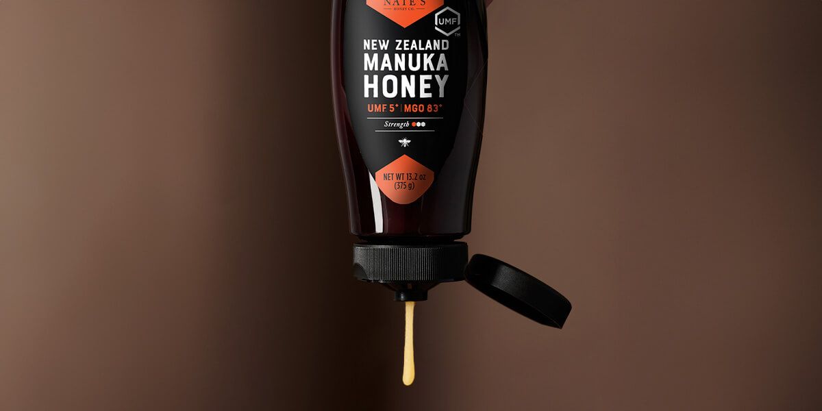 Nate's manuka honey bottle with cap open and honey pouring out. Displayed with brown background.