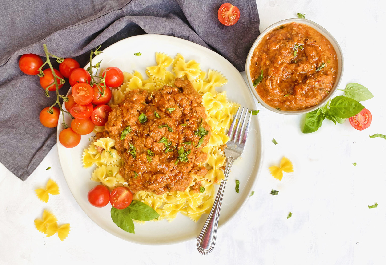 Pasta with sweet potato marinara sauce on plate, cherry tomatoes, basil and grey napkin  on white surface.