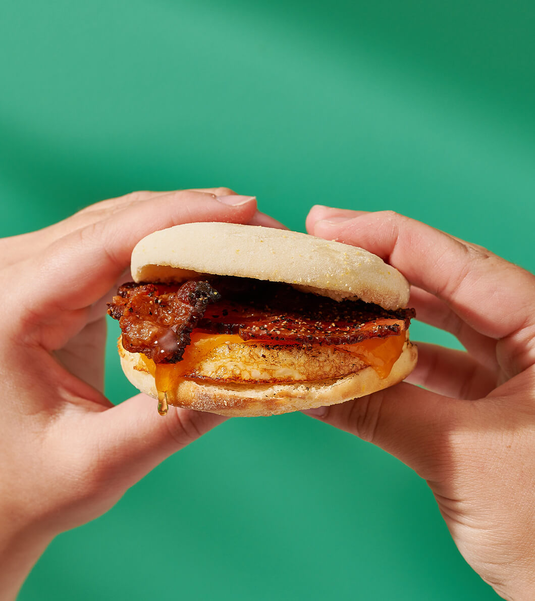 Hands holding a bacon, egg and cheese breakfast sandwich with a drizzle of Nate’s honey against a green background.