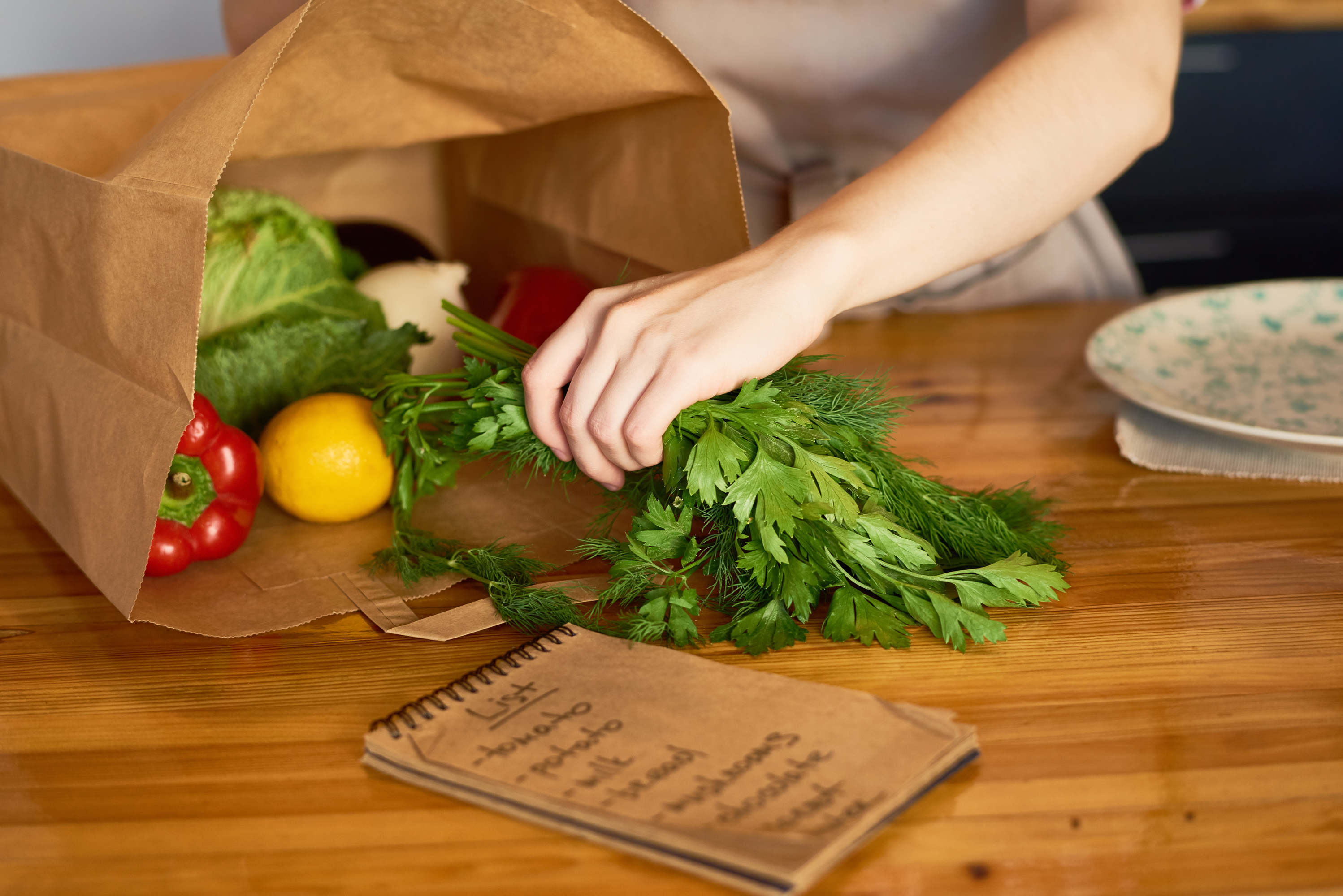 Person unpacking fresh herbs and vegetables with a notebook on a wooden table. Person unpacking fresh herbs and vegetables with a notebook on a wooden table.