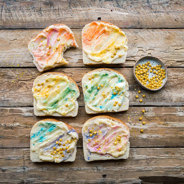 Six slices of bread with colorful spreads and sprinkles on a wooden table, with a bowl of gold candy pearls.