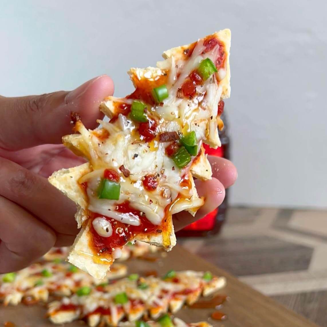 Hand holding mini Christmas tree pizza with hot honey drizzled on top. Minis Christmas tree pizzas and Nate’s honey bottle in background.