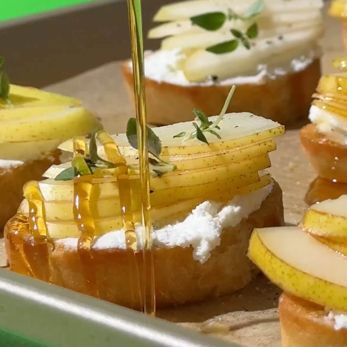 Crostini toast on a pan topped with creamy cheese, pear slices and thyme garnish, and being drizzled with Nate's honey.