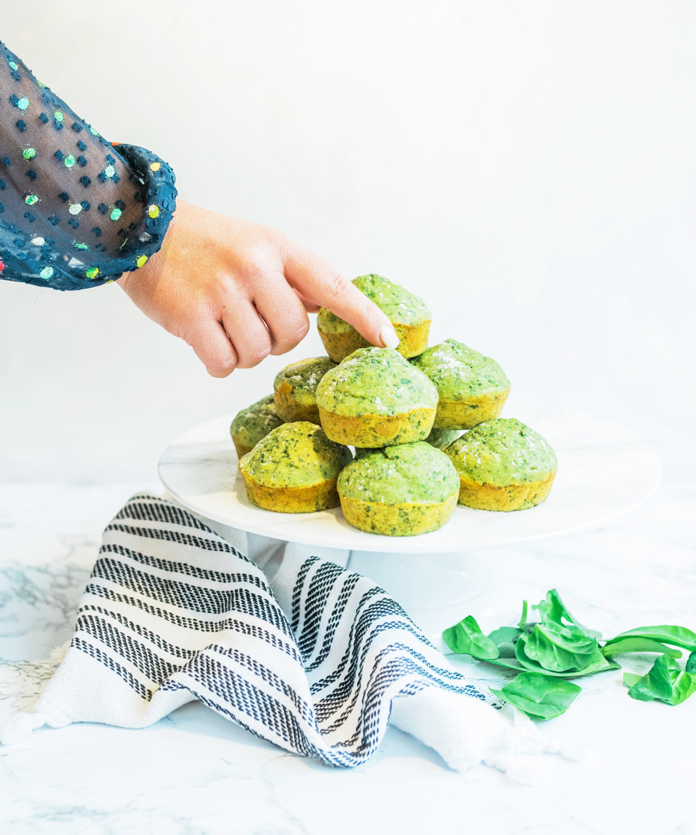 A hand selecting a green spinach and banana muffin from a stacked plate, with spinach leaves and a striped napkin.
