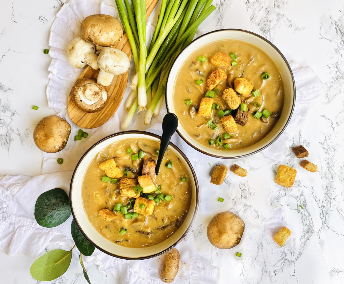 Two bowls of creamy mushroom soup made with Nate’s honey with croutons, herbs and green onions on a marbled background.
