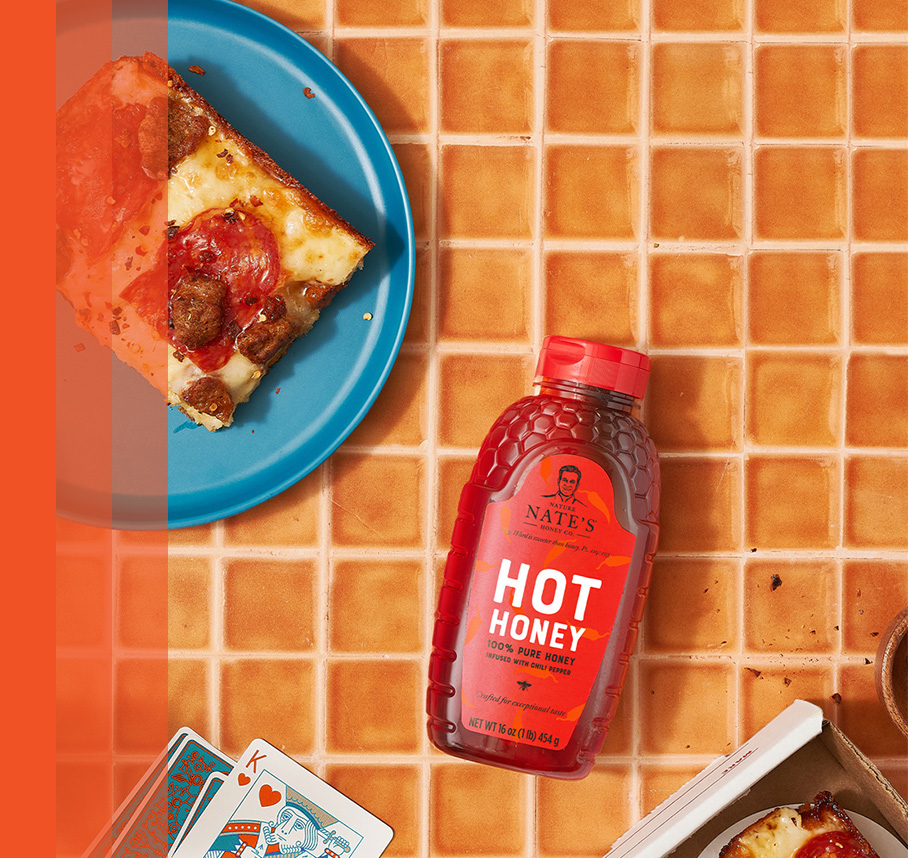 Pizza slices on plates with Nature Nate's Hot Honey, playing cards, on an orange tile table.