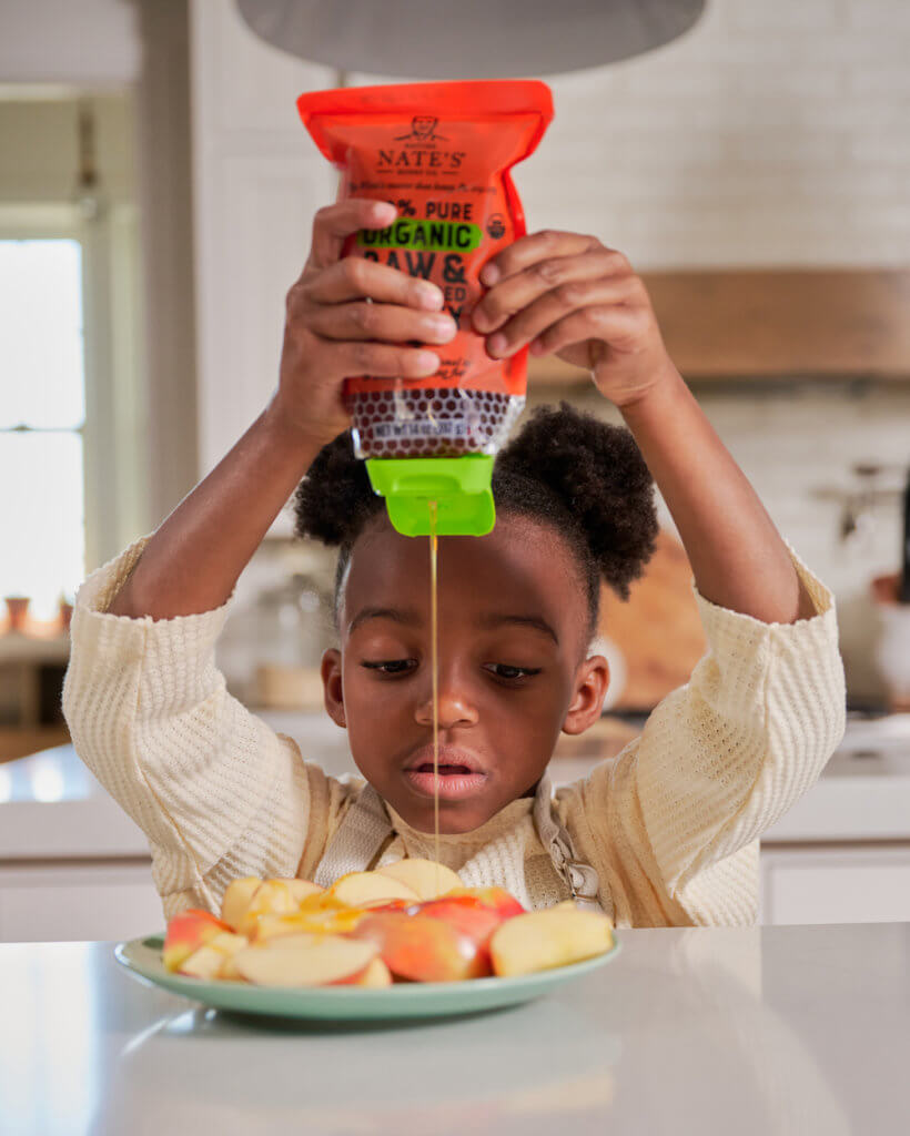 A child focusing intently while squeezing a pouch of Nate's organic honey onto a plate of sliced apples.