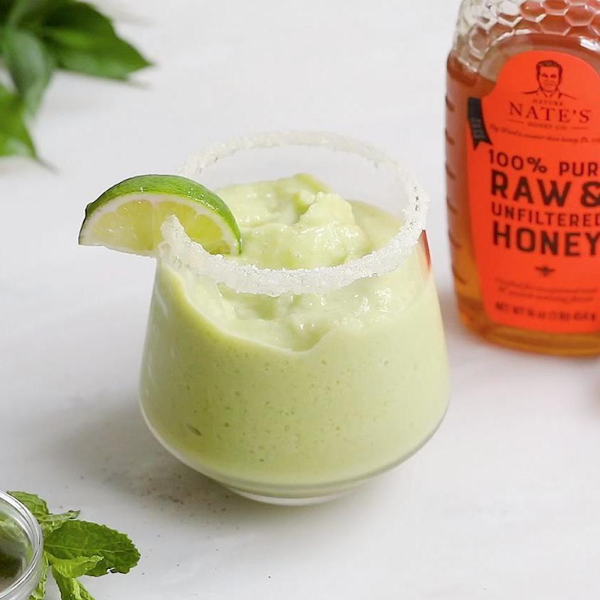 A creamy green smoothie garnished with lime, next to a bottle of Nate’s raw & unfiltered honey.