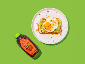A bottle of Nate’s organic honey next to a plate with avocado toast and a sunny-side-up egg on a bright green background.