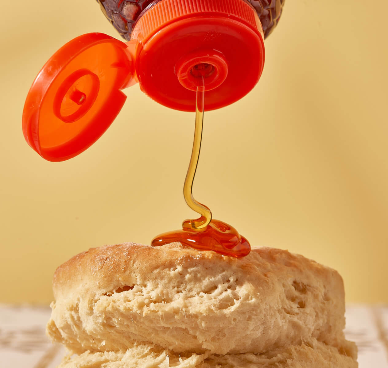 Nate’s honey being drizzled from a bottle onto a fluffy biscuit against a yellow background.