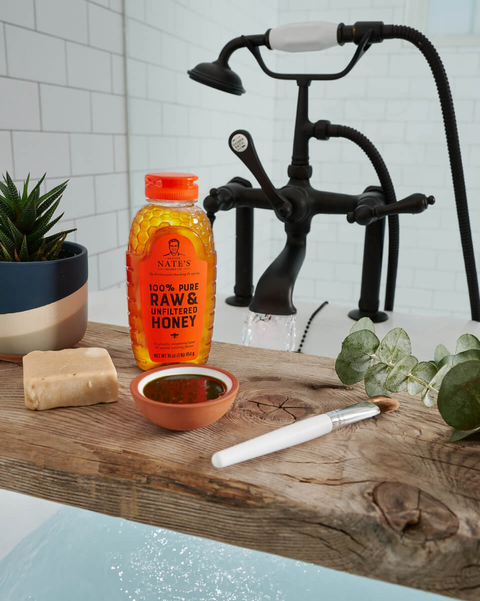 Bottle of Nate's honey, soap, a plant and a white brush on a wood sink counter by a black faucet.