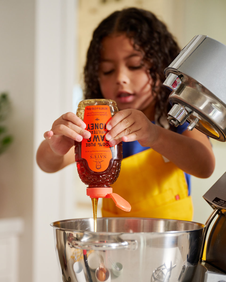 A child in a yellow apron pouring Nate’s honey into a mixing bowl, with a kitchen mixer to the side.
