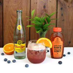 Honey-infused cocktail with oranges, blueberries, sparkling water and a Nate's honey bottle on wooden backdrop.