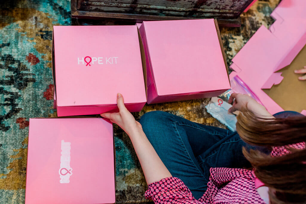 A person sitting on the floor assembling pink boxes labelled, HOPE KIT with a ribbon symbol.