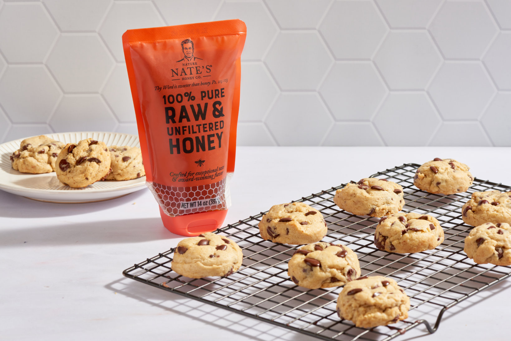 Chocolate chip cookies on a cooling rack and plate, with a pouch of Nate’s 100% pure honey on a kitchen counter.
