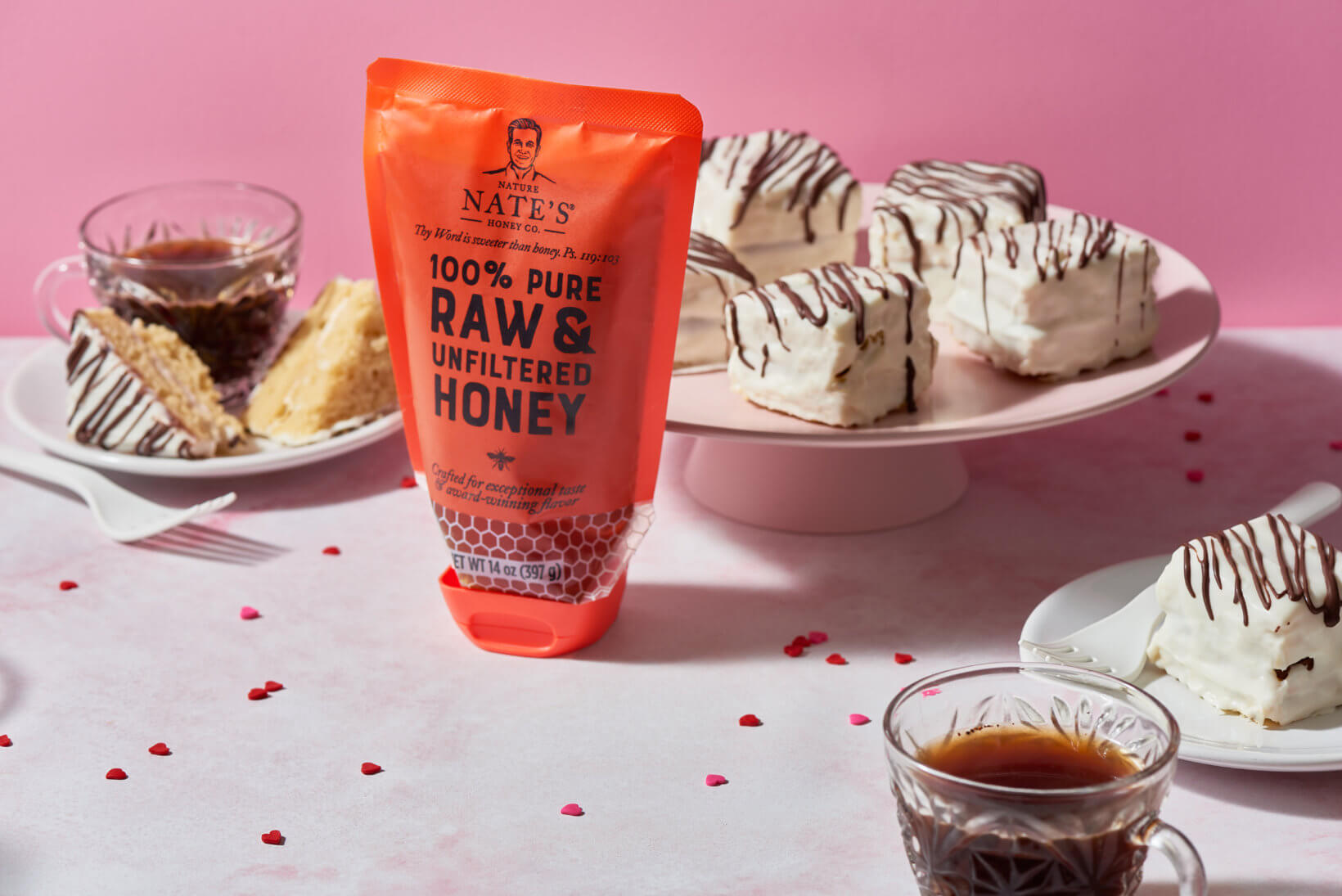 A pouch of Nate's honey, cakes with white icing, and a cup of coffee on a pink background with red sprinkles.