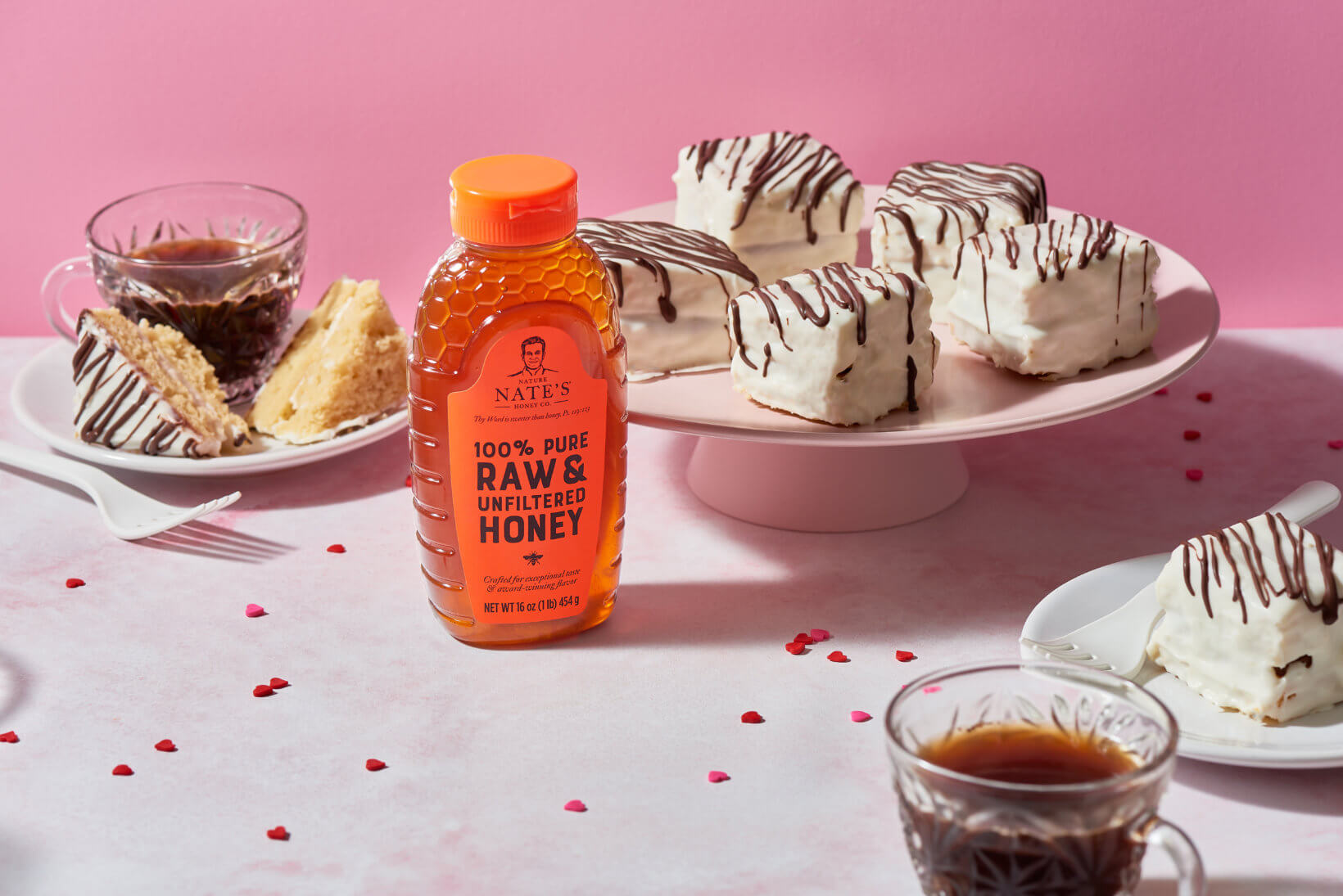 Bottle of Nate's honey, cakes with white icing, and a cup of coffee on a pink background with red sprinkles.