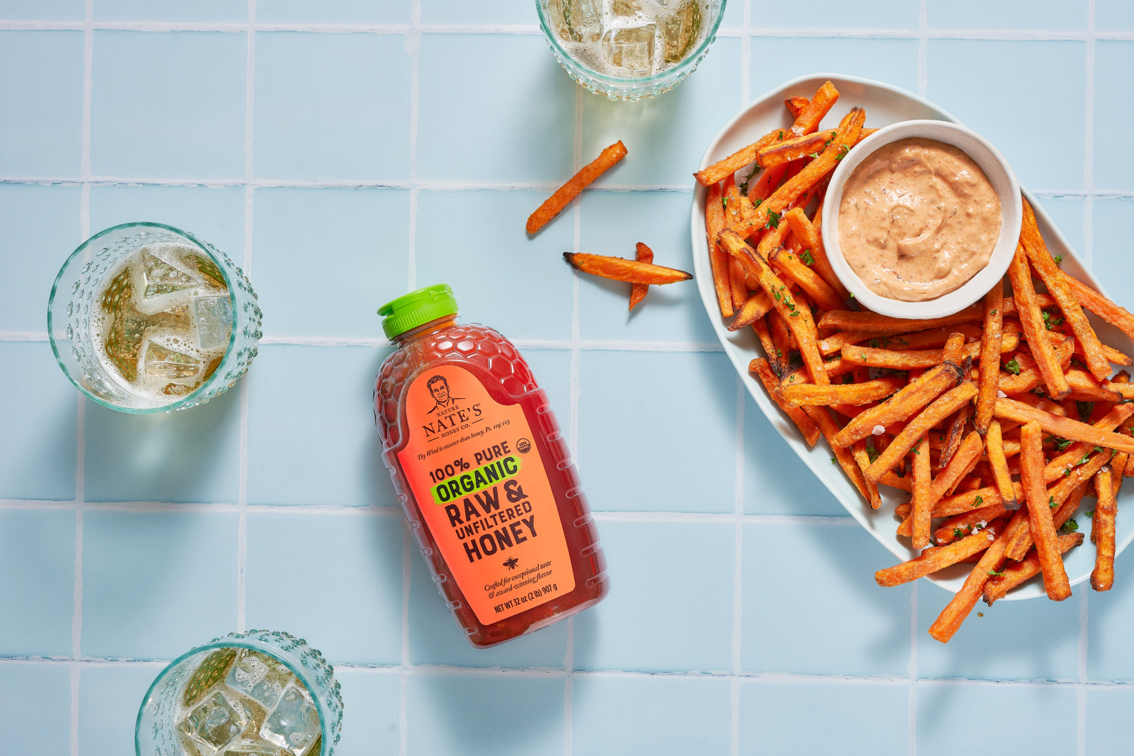Sweet potato fries with dip and a bottle of Nate’s organic honey on blue tiled surface, drinks in background.