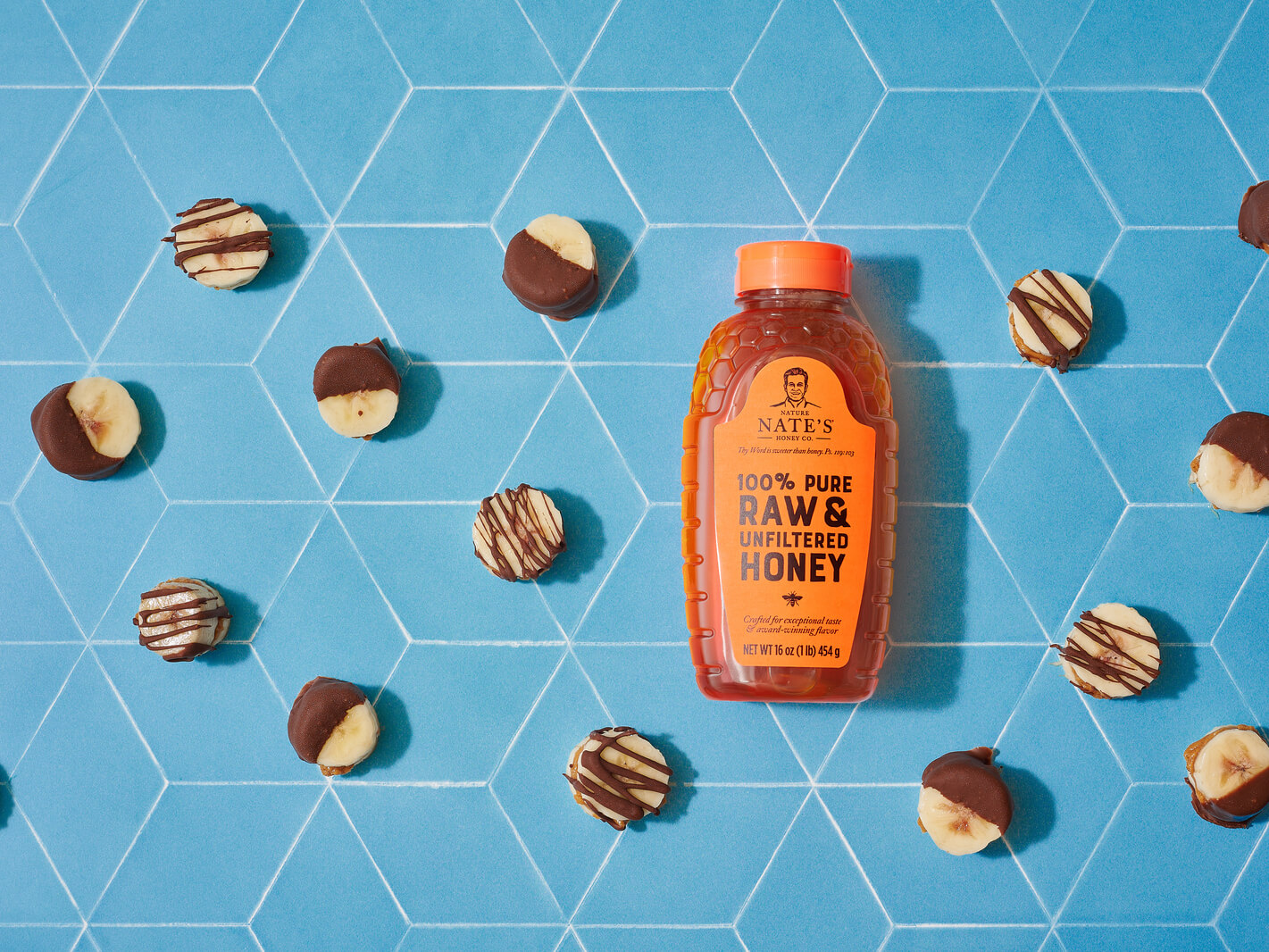 Nate's honey bottle surrounded by chocolate-drizzled banana slices on a blue tile background.