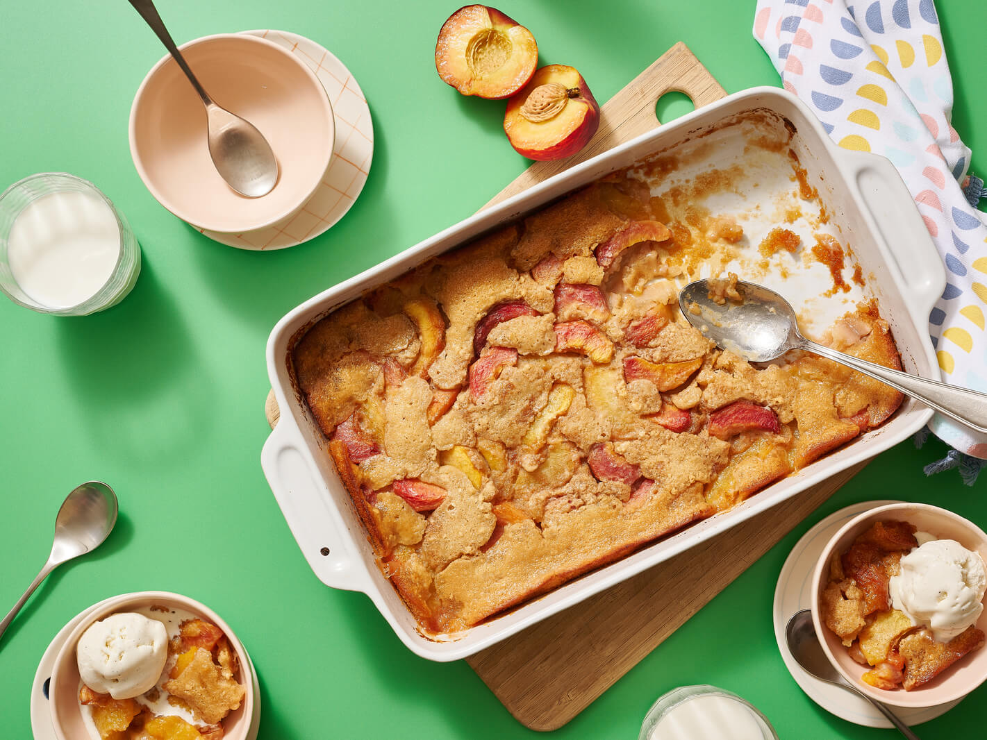 Peach cobbler made with Nate’s honey in a baking dish served with ice cream on a green table with milk and peaches.