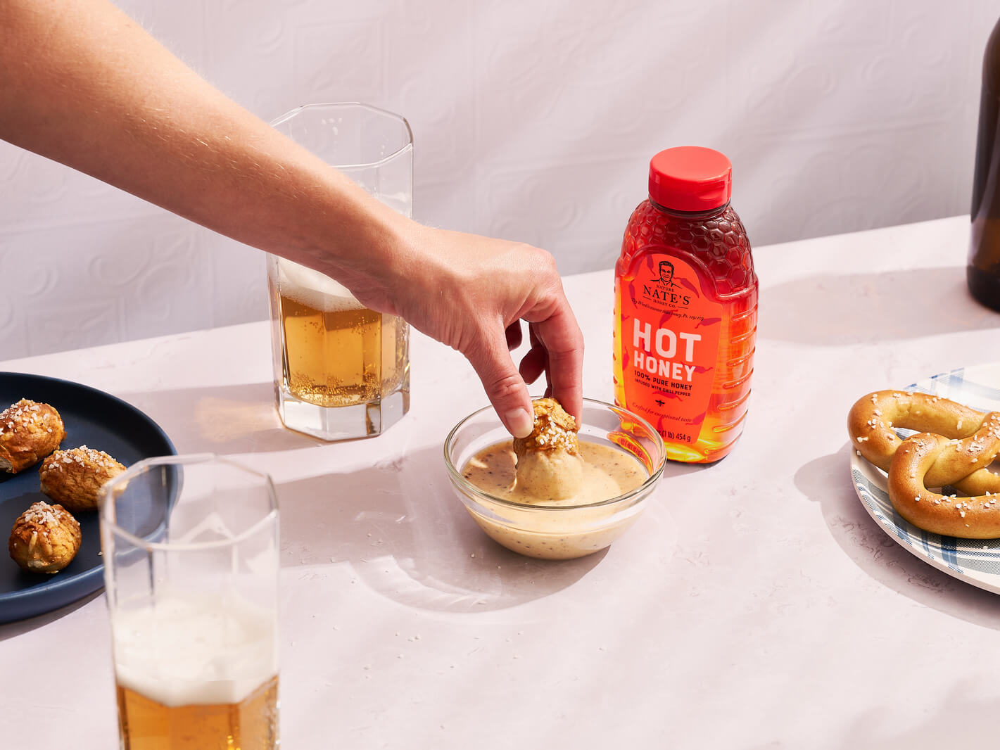 A hand dipping a pretzel in hot honey mustard with a bottle of Nate’s hot honey and drinks in the background.