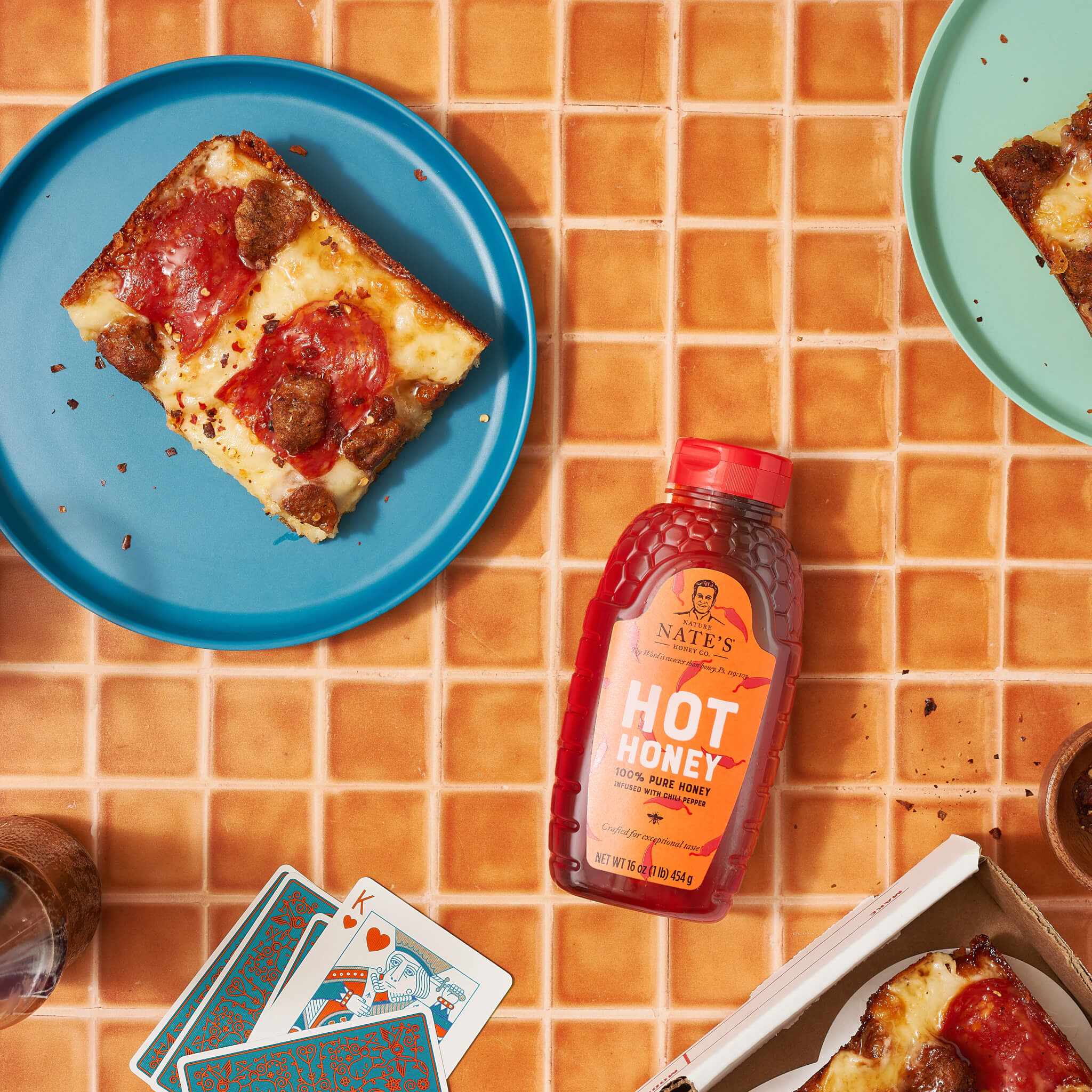 Pizza slices on plates with a bottle of Nate’s hot honey, playing cards and drinks on an orange tile table.
