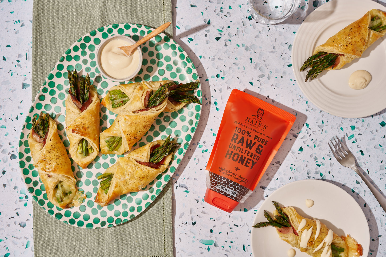 Puff pastry with asparagus, sauce, Nate’s honey pouch, cutlery and water glass on patterned table.