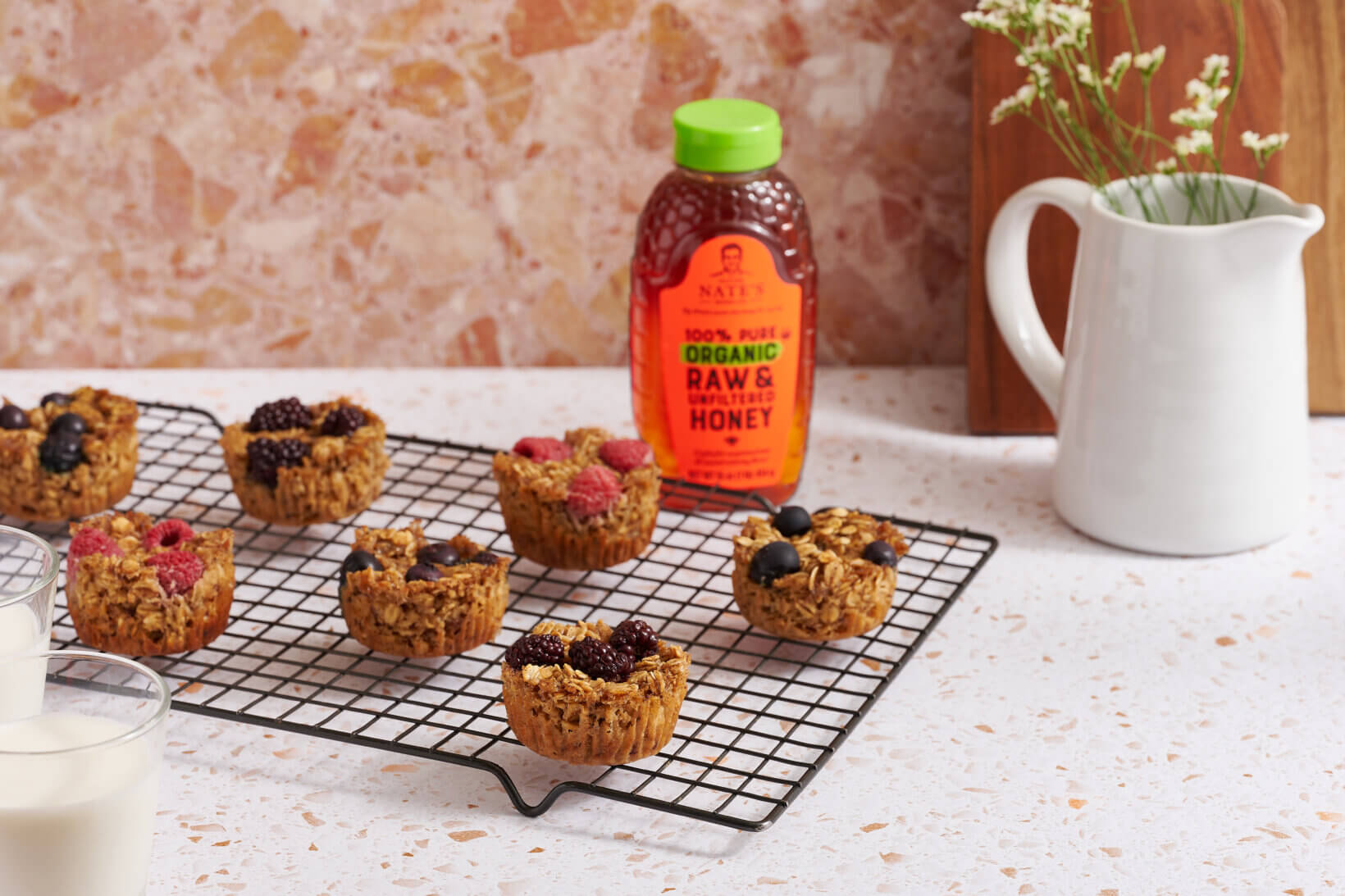 Baked oatmeal cups with berries on a wire rack next to a bottle of Nate’s organic honey and a pitcher of flowers.