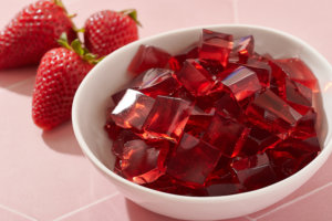 A bowl of red gummy candy cubes made with honey next to fresh strawberries on a pink background.