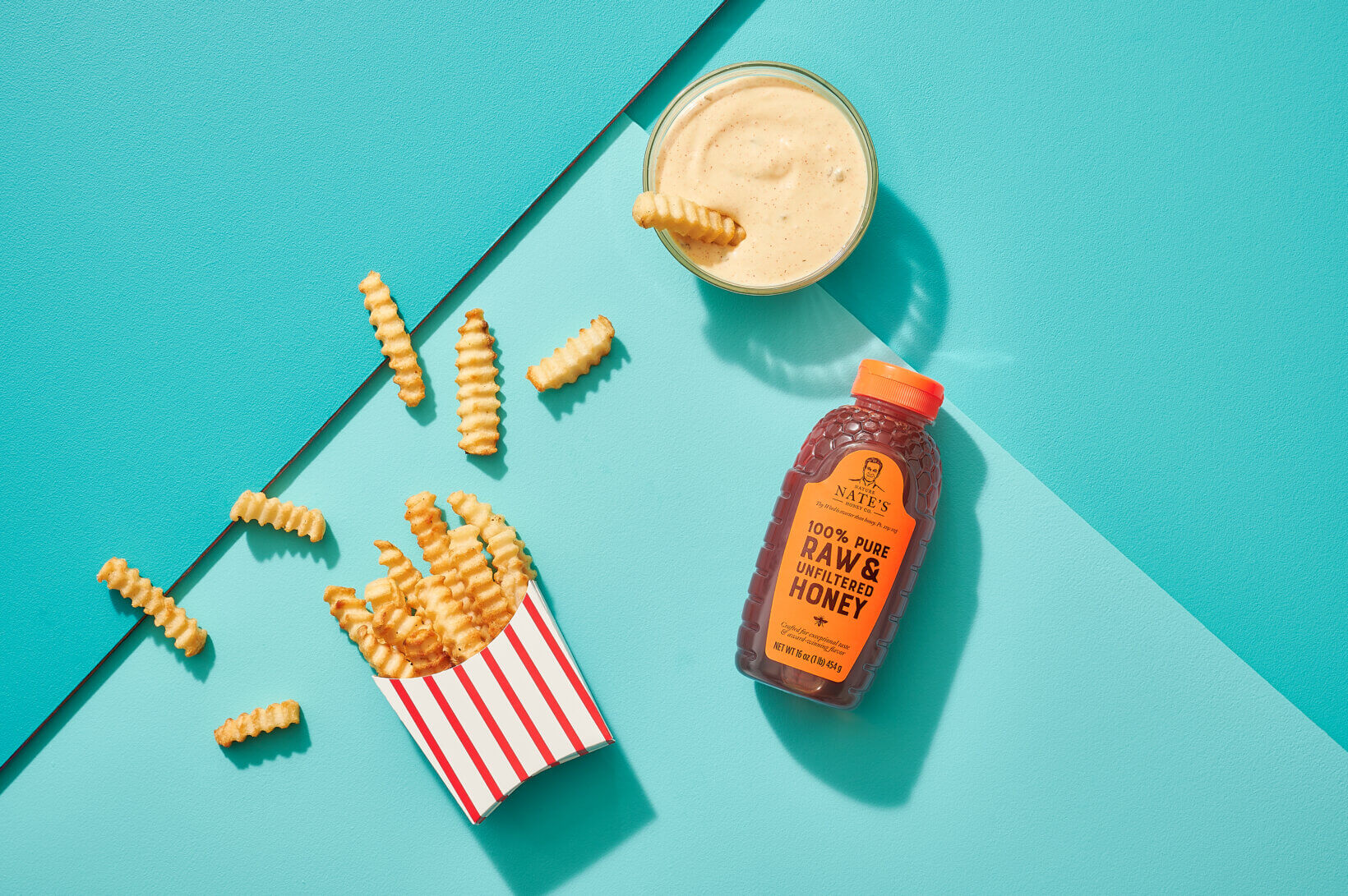 Fries spilling from a striped box with dipping sauce and a bottle of Nate’s honey on a teal background.