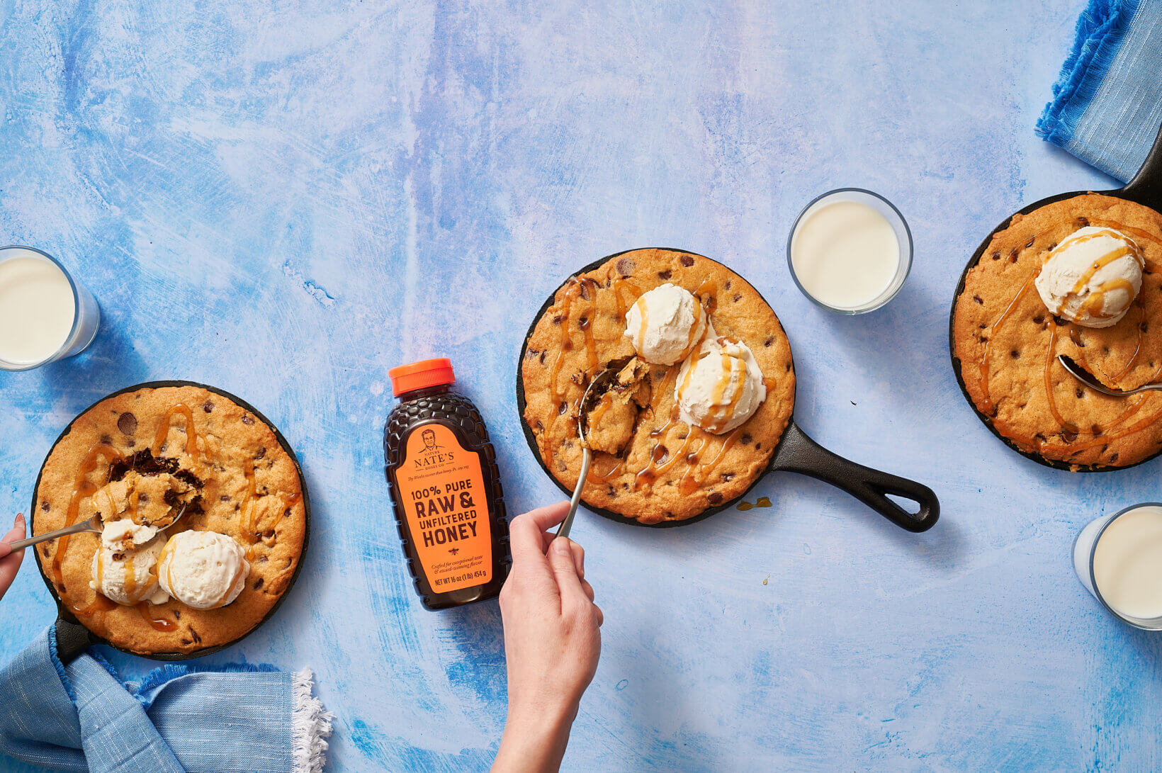 Skillet cookies with ice cream, a bottle of Nate’s honey and glasses of milk with a hand and spoon on a blue background.