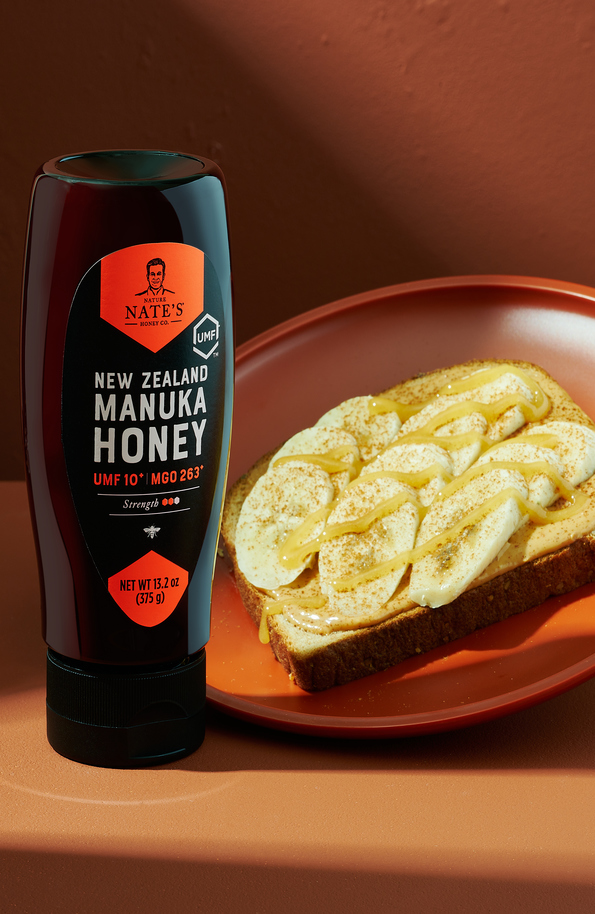 Bottle of Nate's Manuka UMF 10+ next to plate with slice of bread topped with nut butter, banana slices and honey