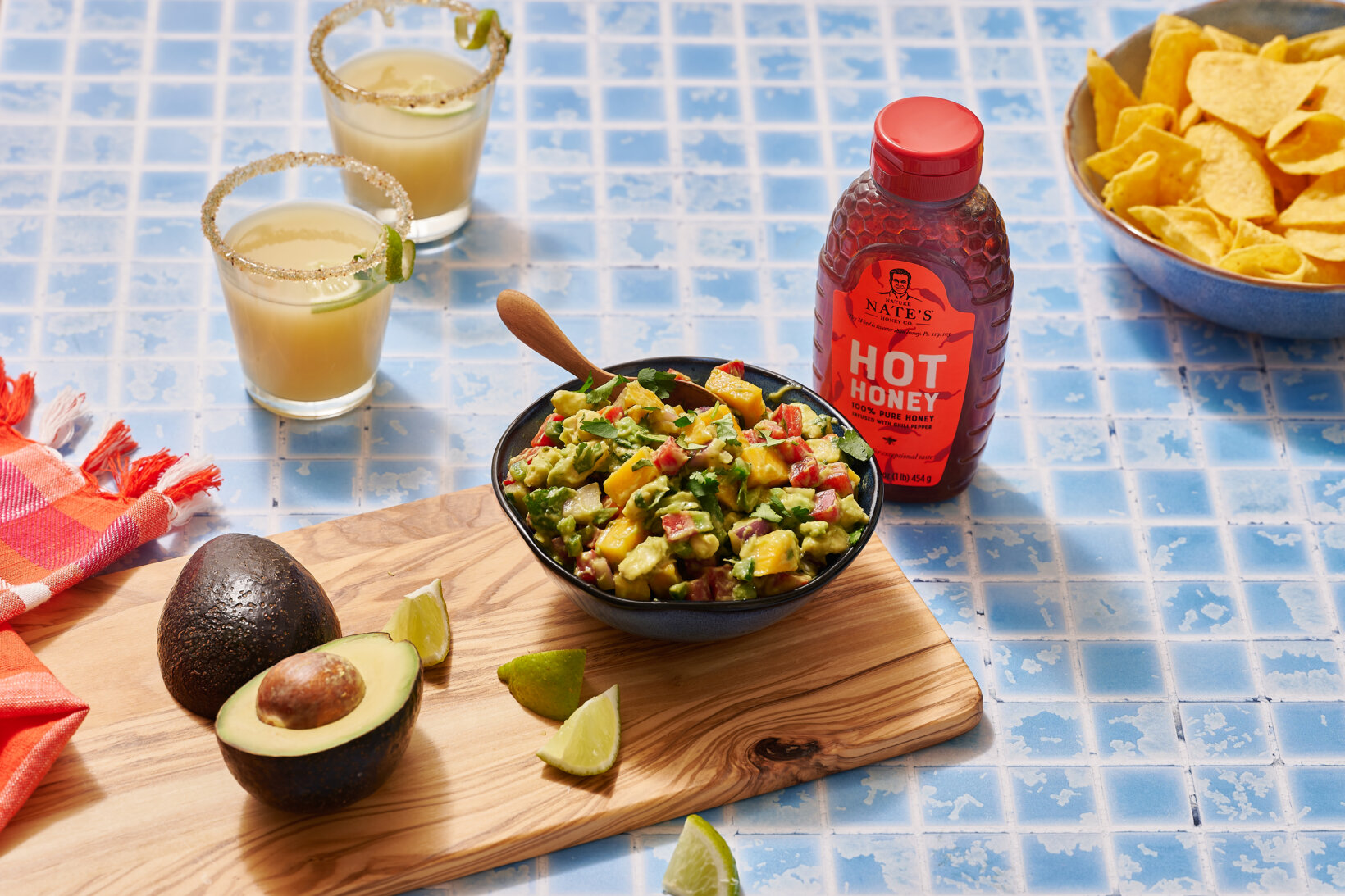 A bowl of mango avocado salsa on a cutting board with avocado, lime, a bottle of Nate’s hot honey, chips and margaritas.