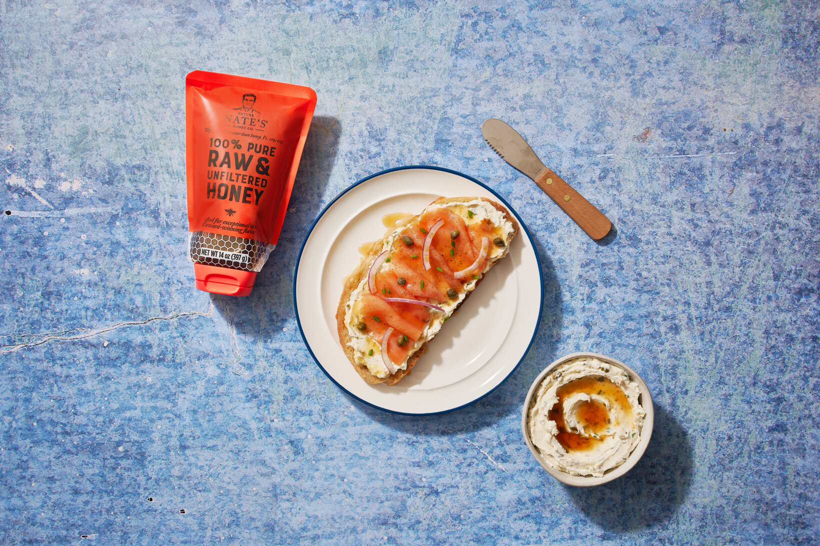 Smoked salmon and cream cheese toast drizzled with honey, next to Nate's honey pouch, knife and bowl on a blue surface.