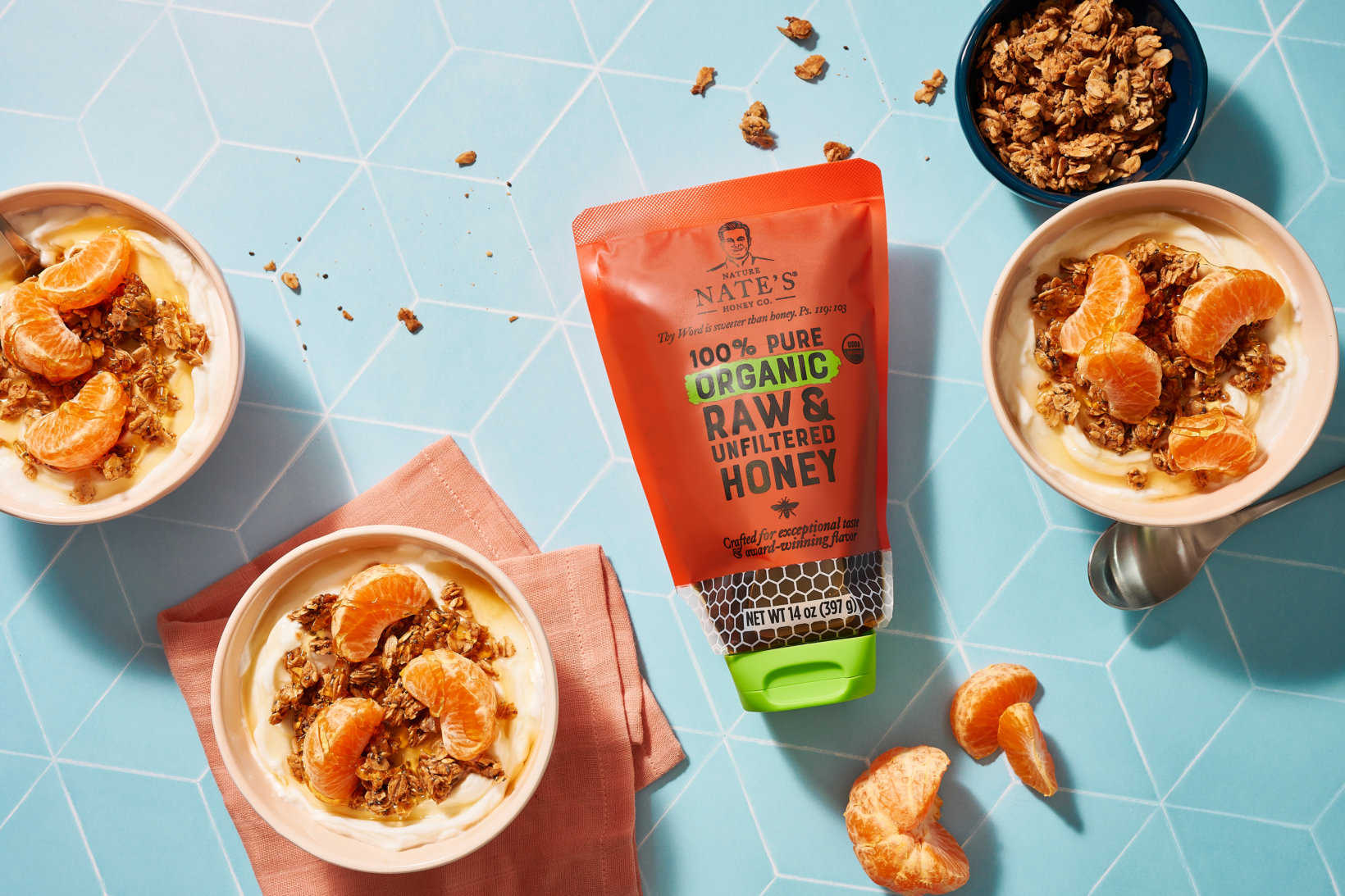 Three bowls of yogurt with orange slices and granola, a pouch of Nate’s honey and granola on a blue-tiled surface.