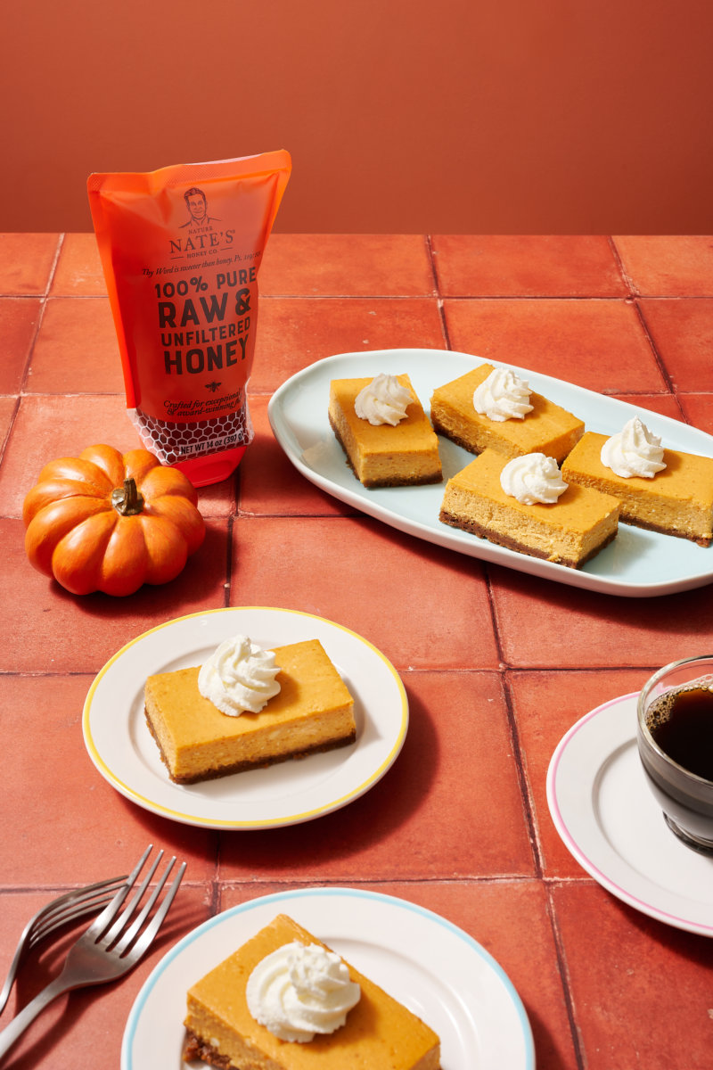 Pumpkin bars on plates, a Nate’s honey pouch, a mini pumpkin, cutlery and a cup of coffee on a terracotta tiled table.
