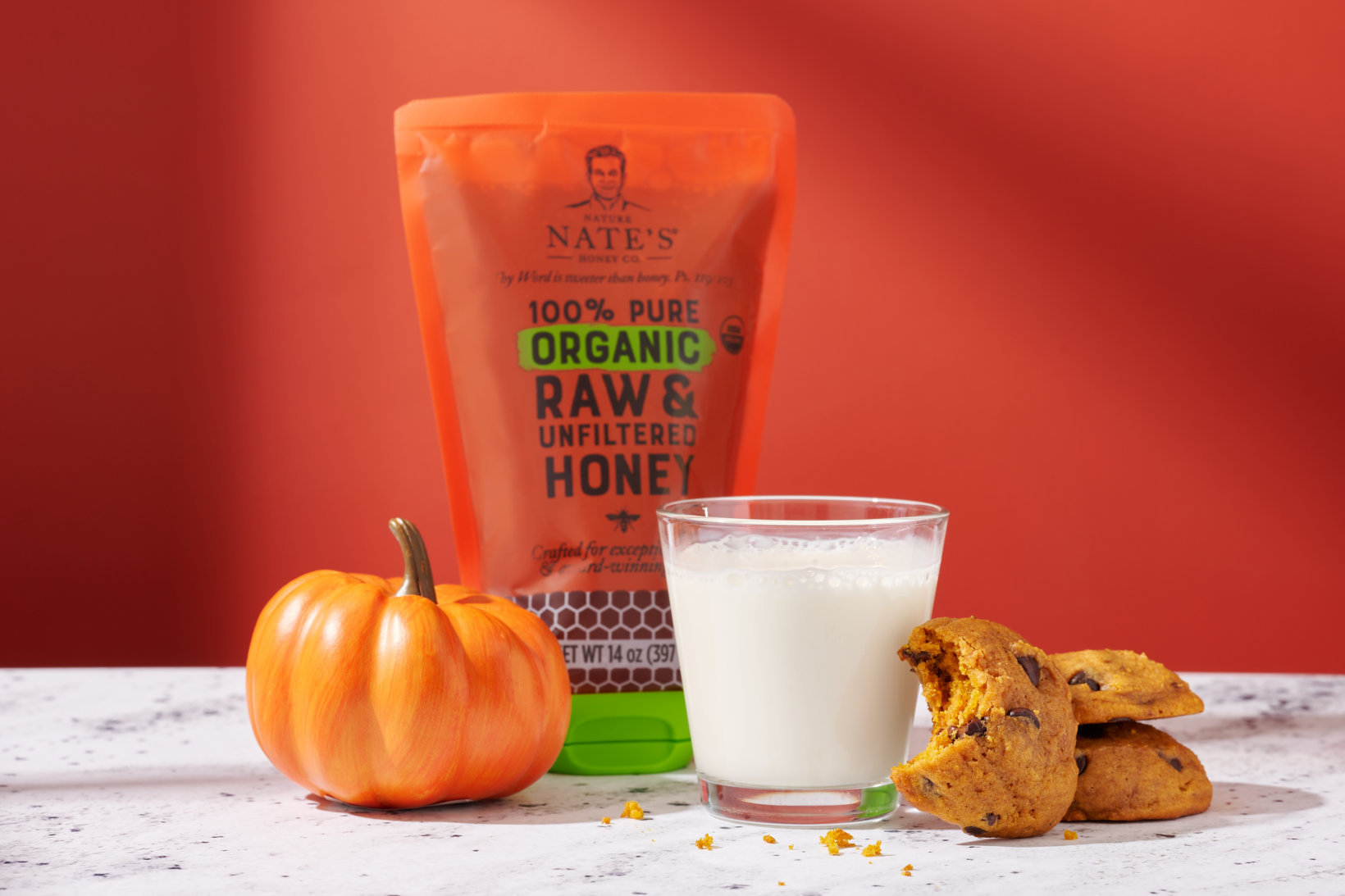 A festive setting with a Nate's organic honey pouch, a glass of milk and pumpkin cookies with a warm backdrop.
