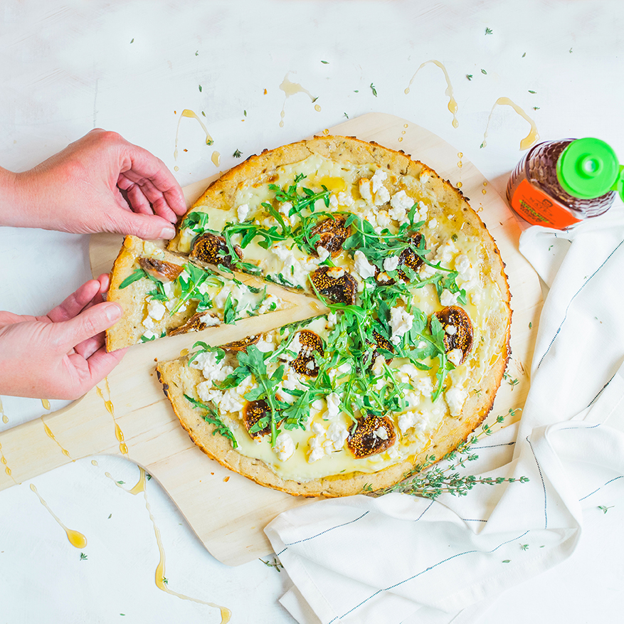 Hands serving a slice of mushroom and herb pizza on a cutting board, with a bottle of Nate’s organic honey.