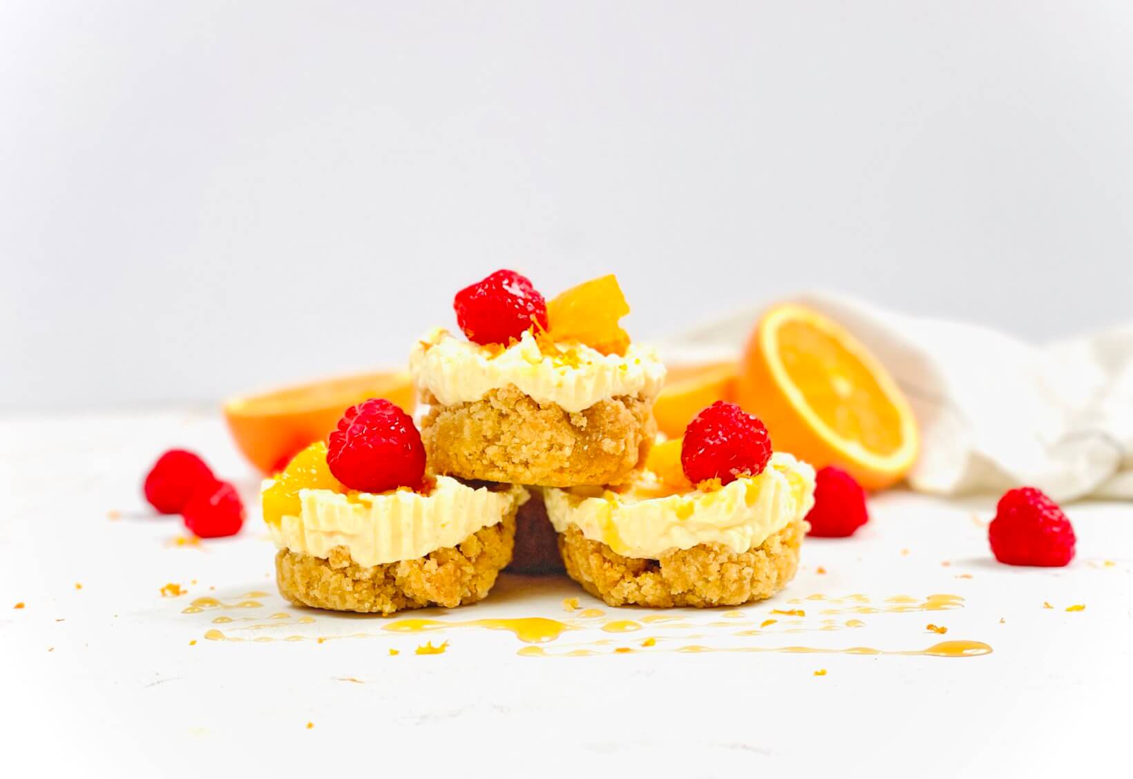 Stacked cheesecake bites, garnished with honey, fresh  raspberries and orange pieces, against a clean background.