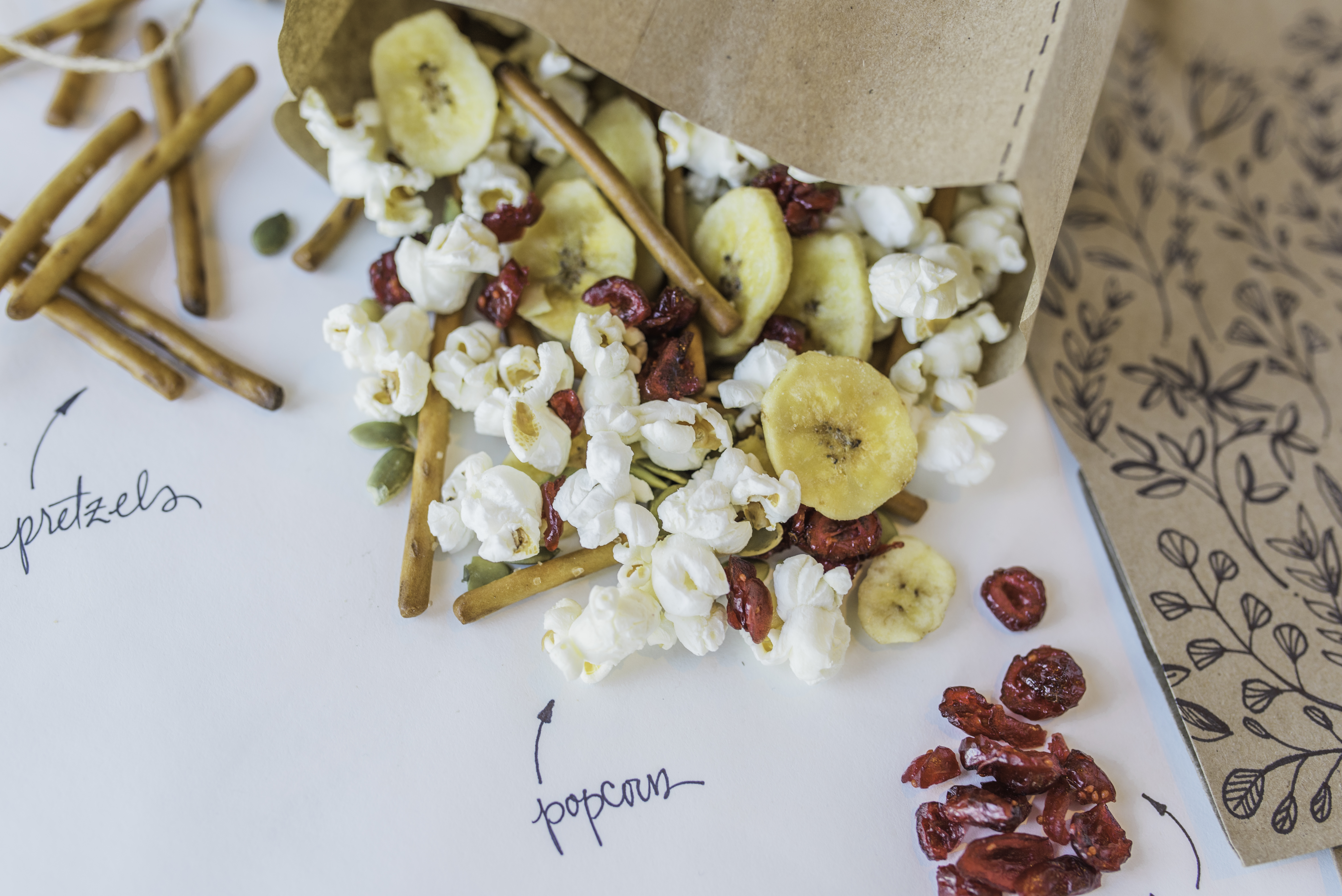 Popcorn, pretzels, seeds, dried bananas and cranberries spilling from paper bag on a labelled surface.