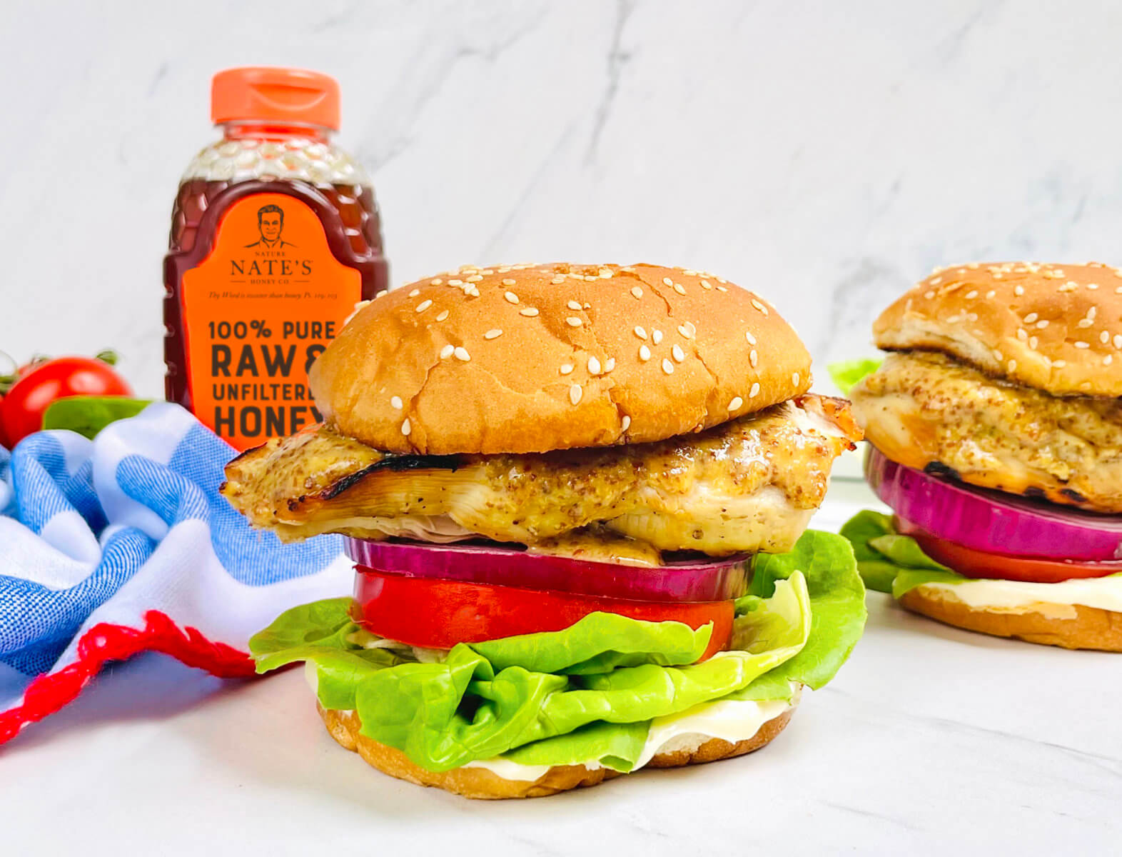 Hot honey mustard chicken sandwiches with lettuce and tomato, and a bottle of Nate’s honey on a marble surface.