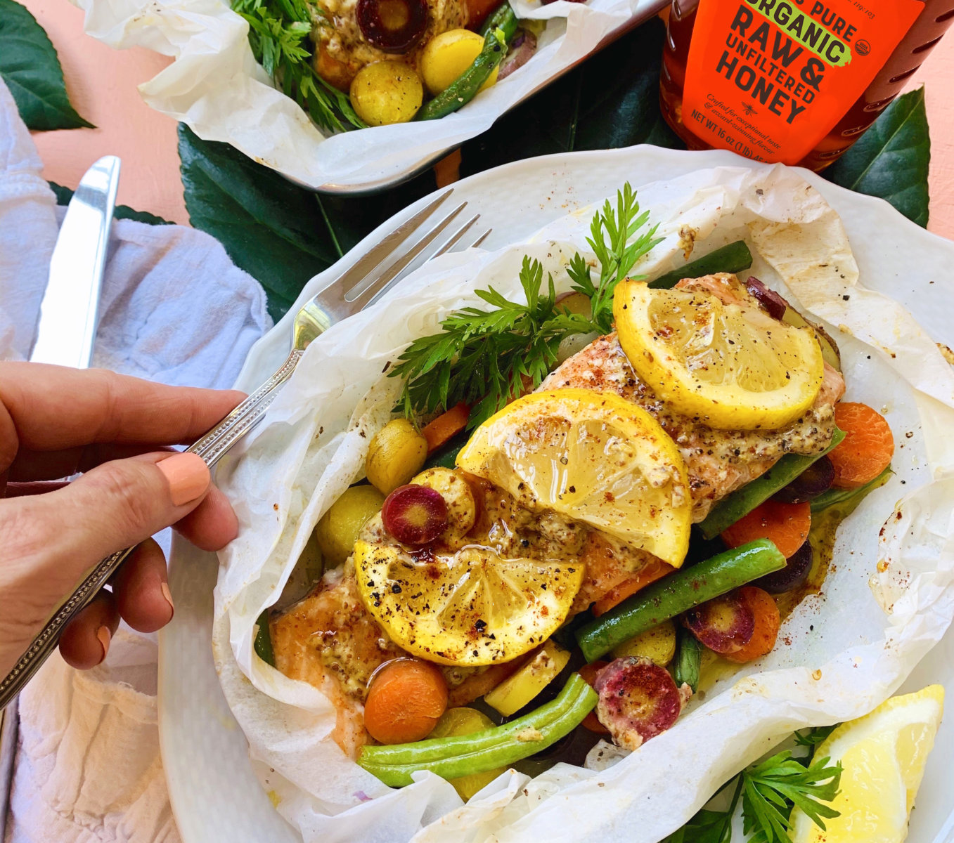 Hand holding a fork with baked salmon with vegetables and lemon in parchment paper, and a bottle of Nate's honey nearby.