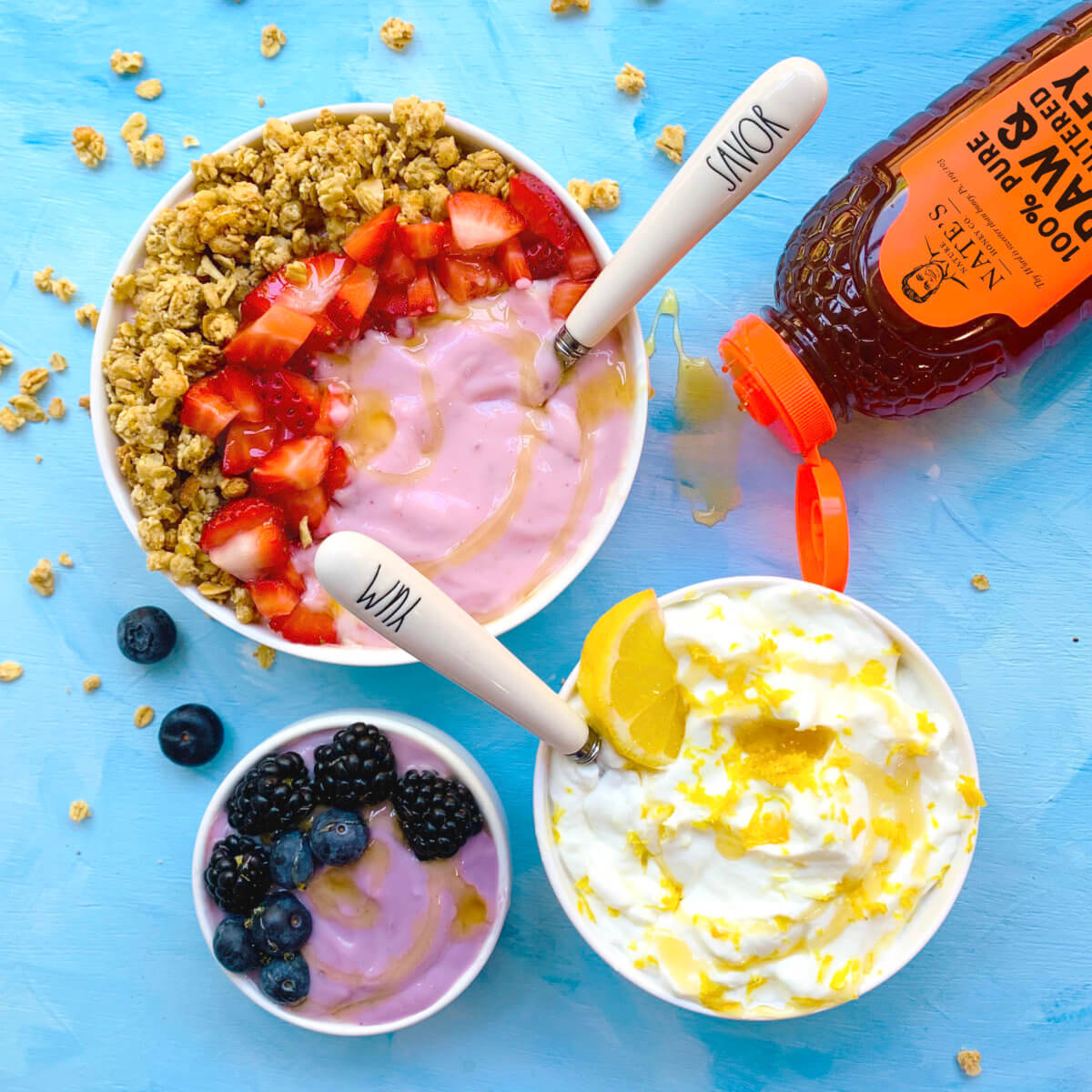 Yogurt bowls with granola, berries and honey drizzle with a Nate's honey bottle on a blue surface with spoons.