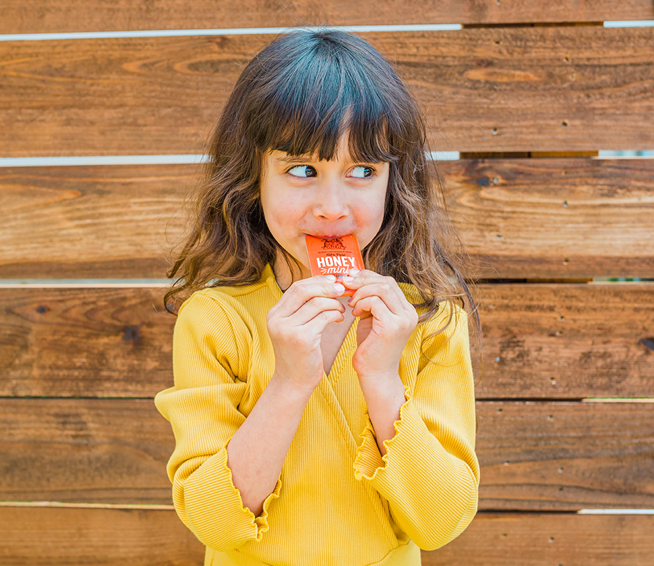 Child in yellow, eating a Nate's honey mini, against a wooden backdrop.