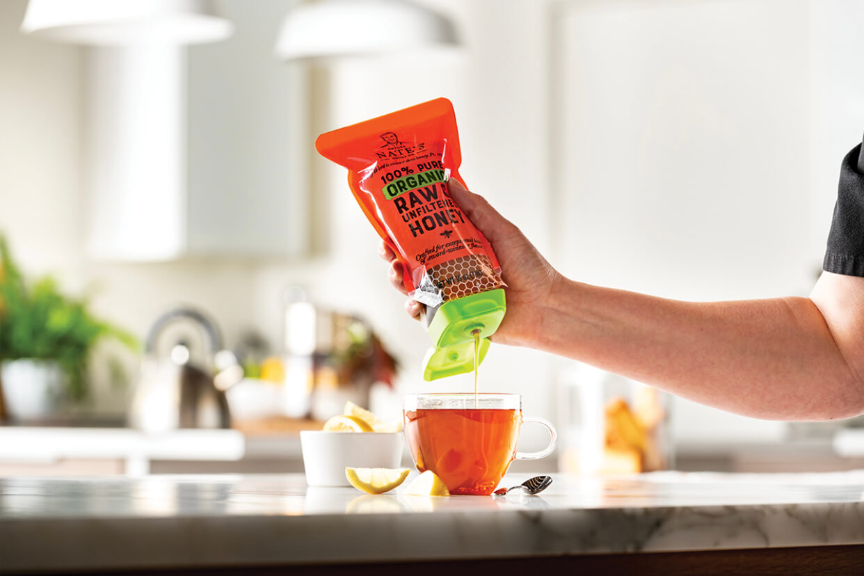Person pours organic honey into a cup of tea, with lemon slices nearby in a kitchen setting.